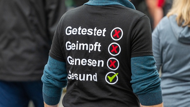 Mann auf einer Demonstration mit schwarzem T-Shirt mit Aufdruck: Genesen, Geimpft, Genesen, Gesund. Nur Gesund ist angehakt. | Bild: picture alliance
