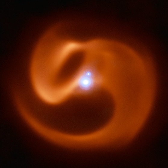 Infrarot-Aufnahme des Sternsystems Apep in der Konstellation Winkelmaß. Es besteht aus einem doppelten Wolf-Rayet Stern sowie einem Begleitstern, die in der Mitte des Bildes hell leuchten. Sie sind von einer rötlichen Spirale aus Gas und Staub umgeben, die von dem Wolf-Rayet Stern in der Mitte erzeugt werden. | Bild: ESO