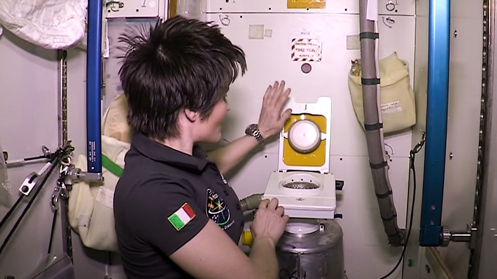 Die italienische Astronautin Samantha Cristoforetti zeigt die ISS-Toilette. Rechts an der Wand hängt der Urinschlauch. | Bild: ESA auf Youtube