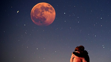 Sterngucker beobachten eine totale Mondfinsternis: Ein Pärchen betrachtet eng umschlungen den abendlichen Sternenhimmel voller Sterne und dem Blutmond bei einer MoFi (Collage) | Bild: pa/dpa; Montage: BR