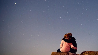 Sterngucker: Ein Pärchen betrachtet eng umschlungen den abendlichen Sternenhimmel voller Sterne.  | Bild: picture-alliance/dpa