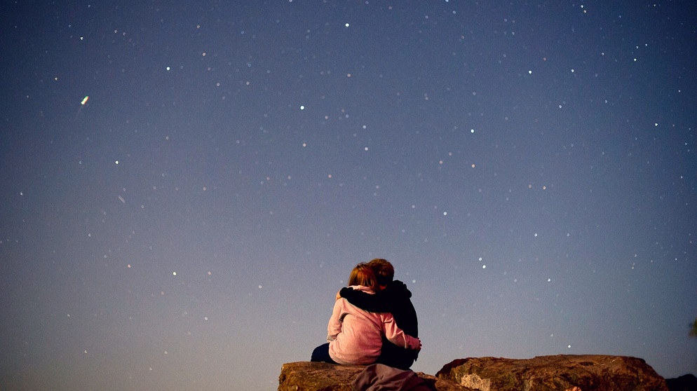 Sterngucker: Ein Pärchen betrachtet eng umschlungen den abendlichen Sternenhimmel voller Sterne.  | Bild: picture-alliance/dpa